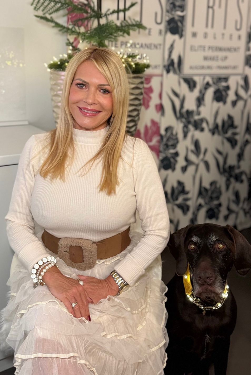 Iris Wolter in cremefarbenem Pullover und weißem Rock sitzt drinnen neben einem schwarzen Hund und lächelt in die Kamera.