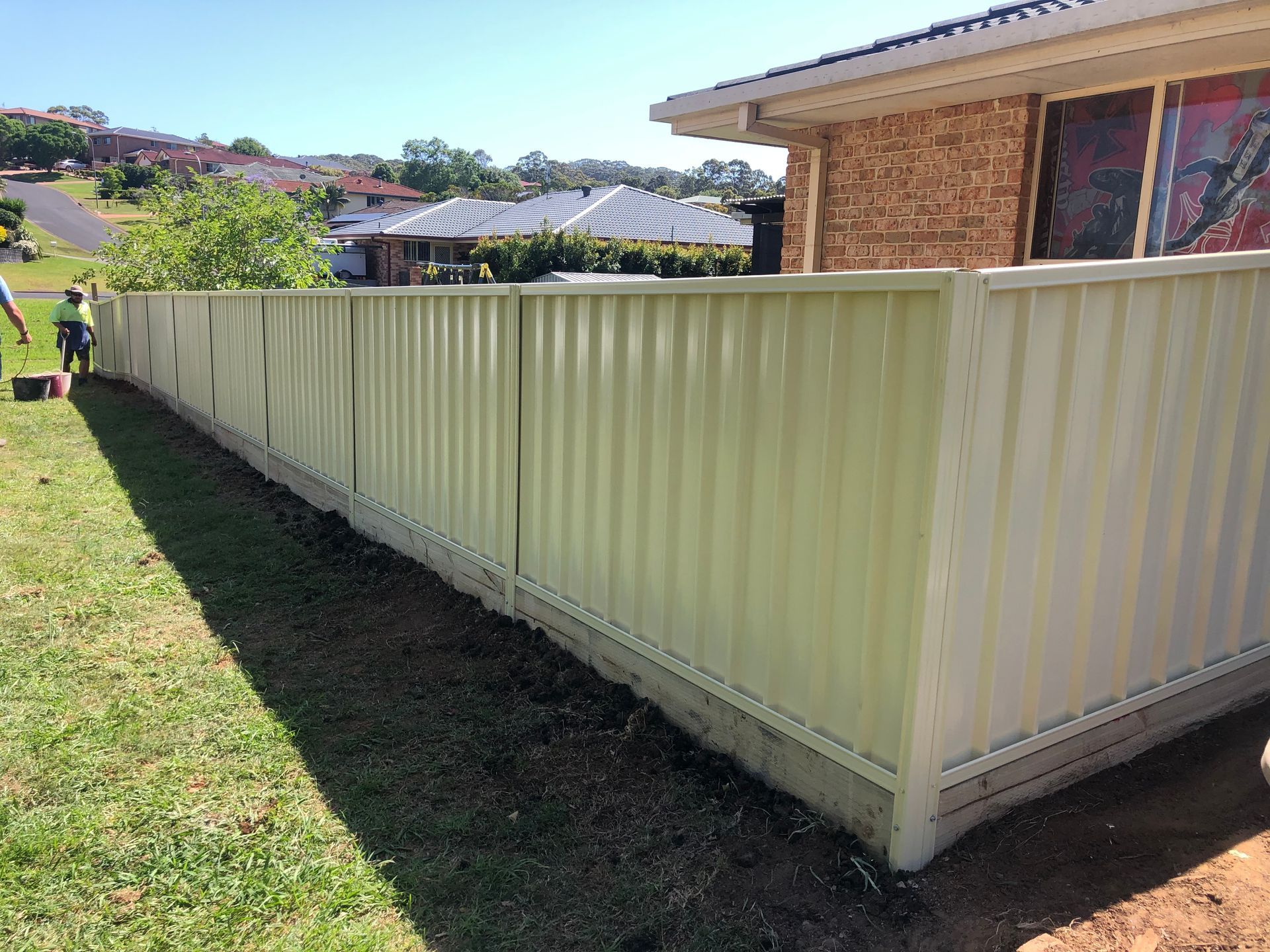 Tan corrugated metal fence bordering a grassy yard, next to a brick house, with a row of houses in the background — Fencer Steve In Temagog, NSW