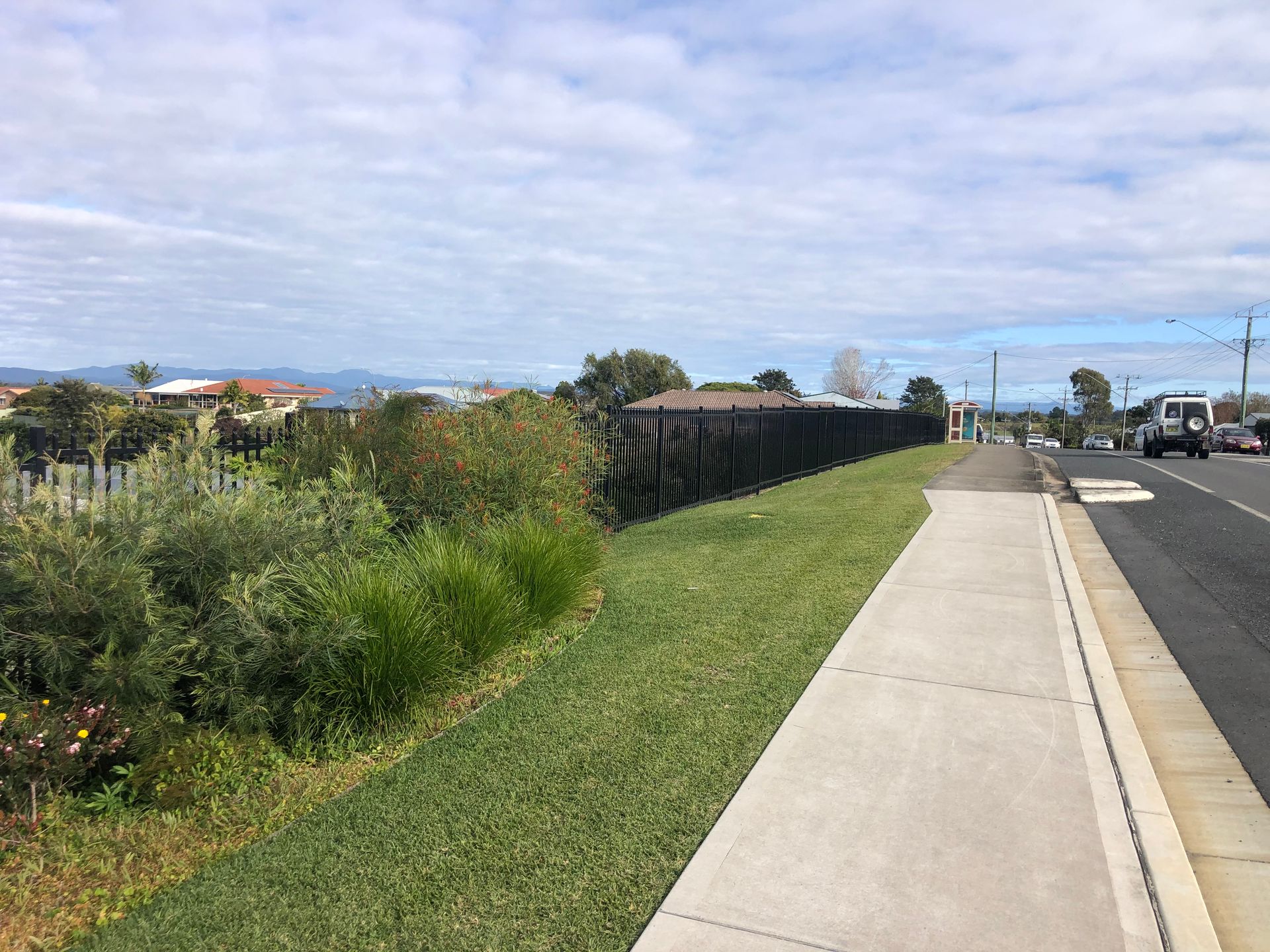 Sidewalk next to a road with grass and a black fence; cloudy sky in background — Fencer Steve In Temagog, NSW