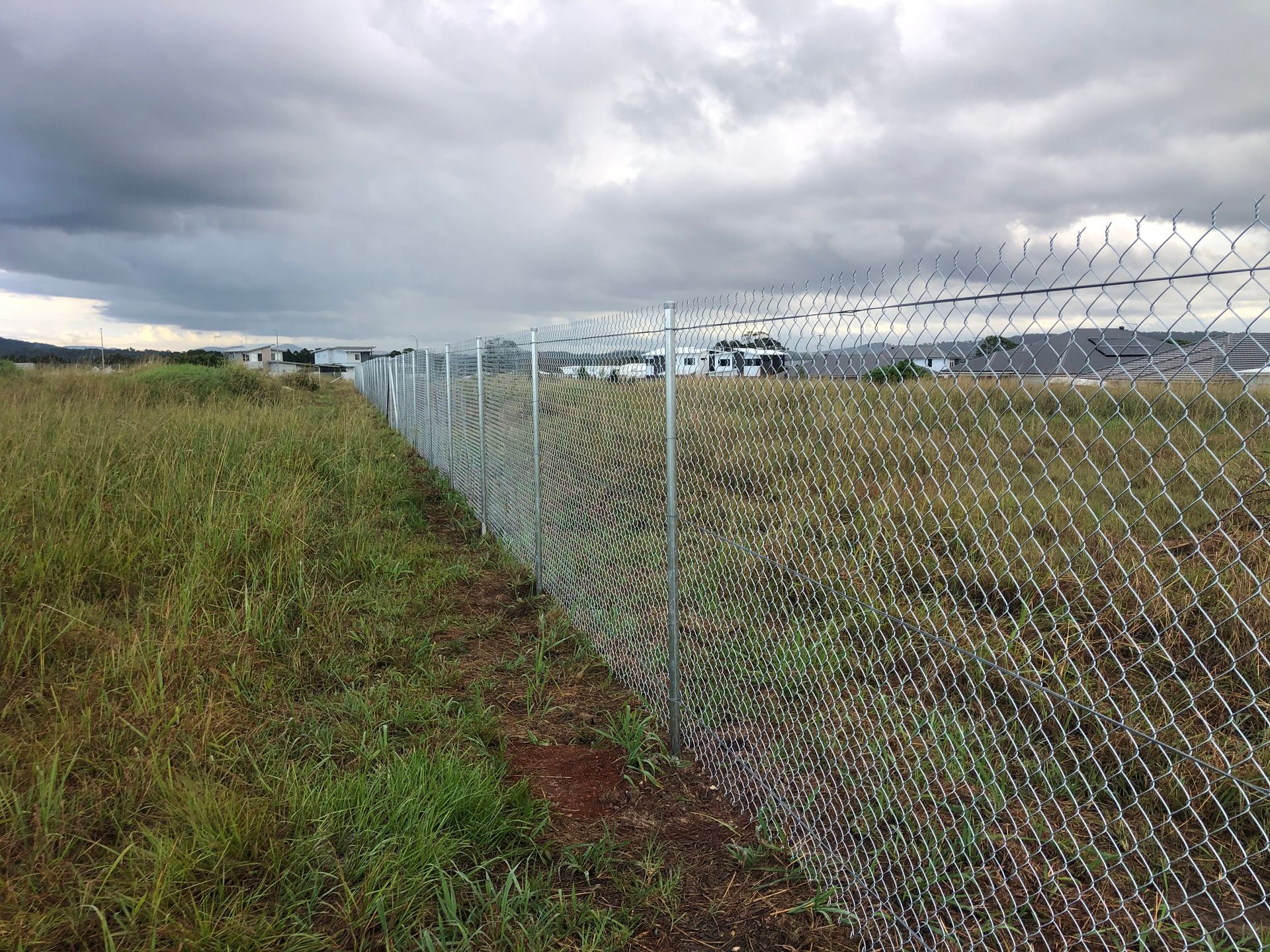 Chain-link fence bordering a grassy field, with houses visible in the distance under a cloudy sky — Fencer Steve In Temagog, NSW