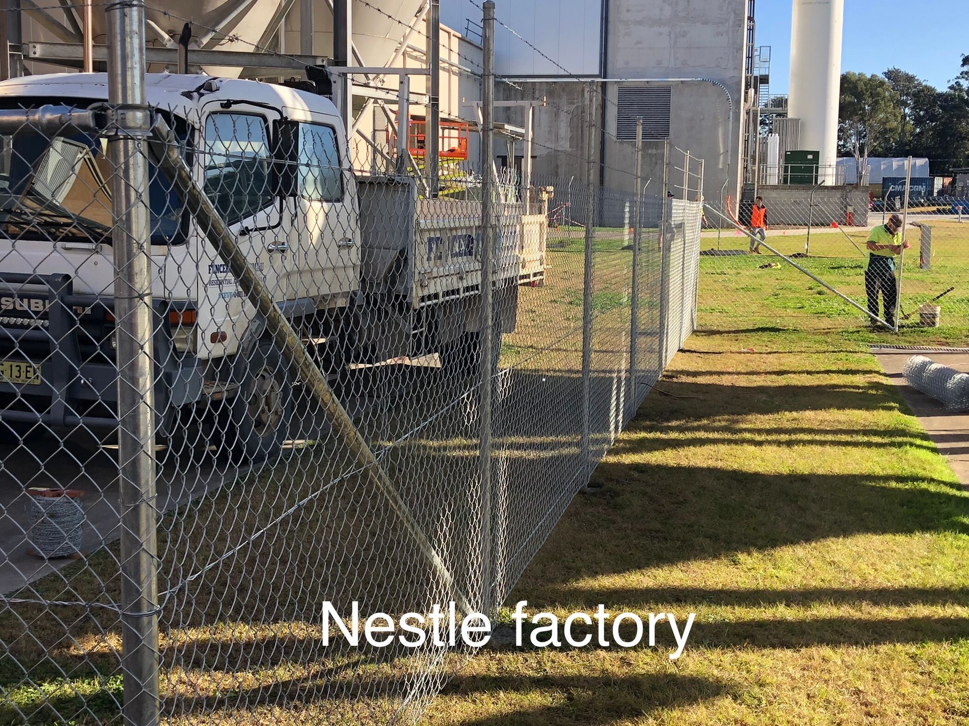 Chain-link fence surrounding a Nestlé factory; a truck and worker mowing grass are visible — Fencer Steve In Temagog, NSW