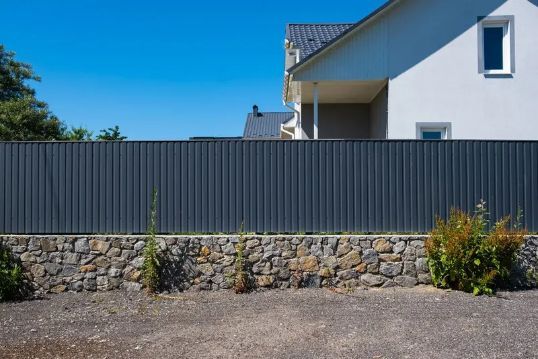 Gray Metal Fence Atop Stone Wall, With a House and Clear Sky in the Background — Fencer Steve In South West Rocks, NSW