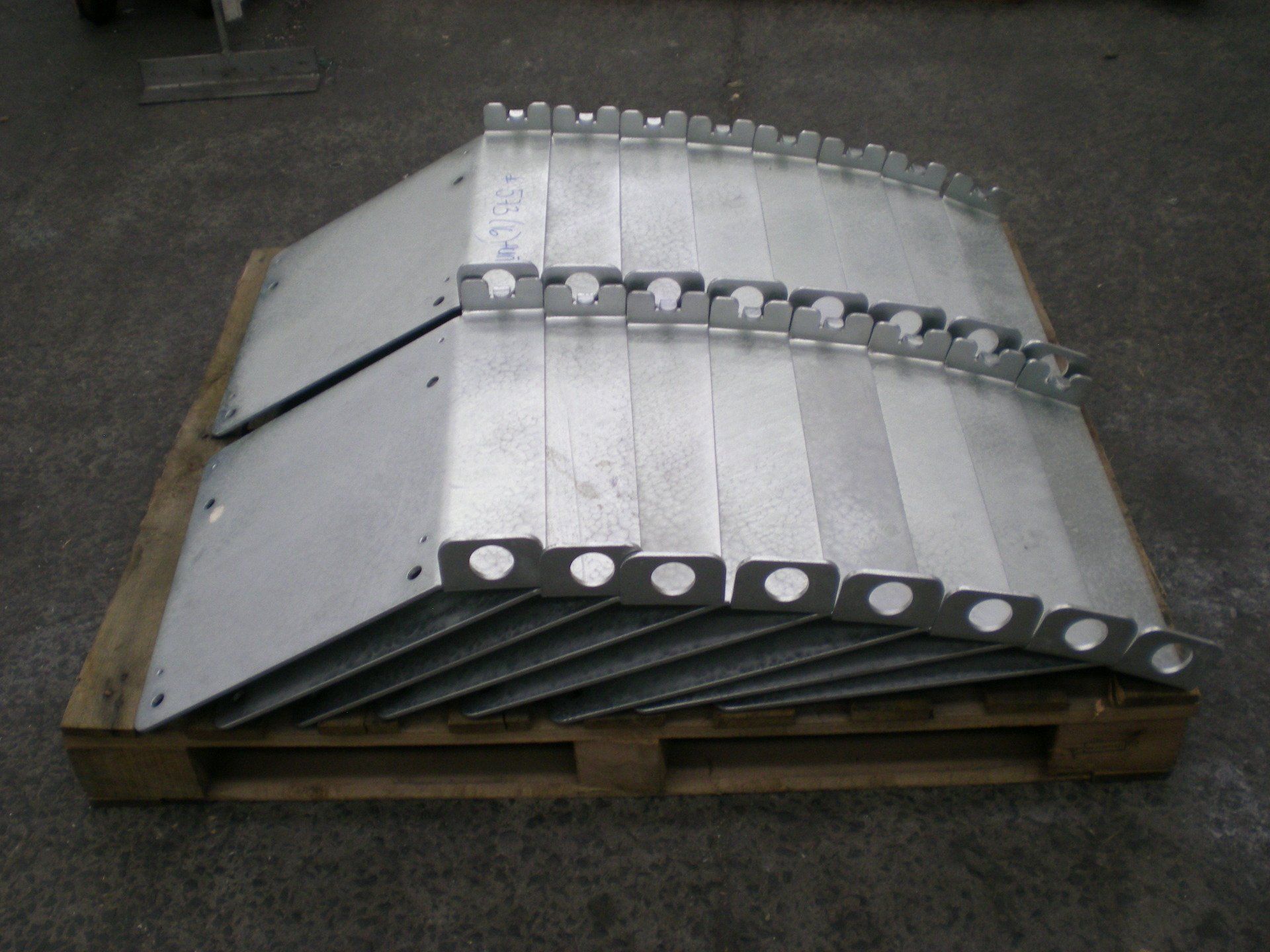 Metal plates stacked on a wooden pallet. The plates are silver, with holes along one edge, and rest on a concrete floor.