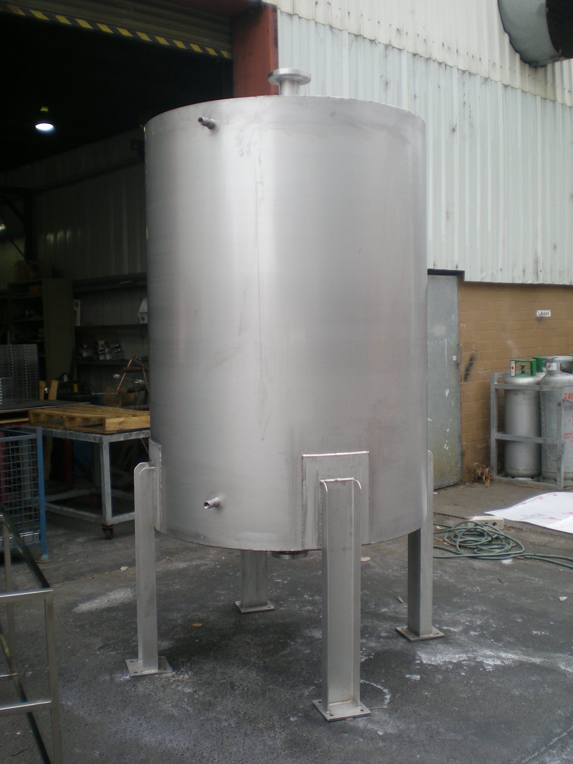 A tall, stainless steel industrial tank with supporting legs sits in a factory setting.