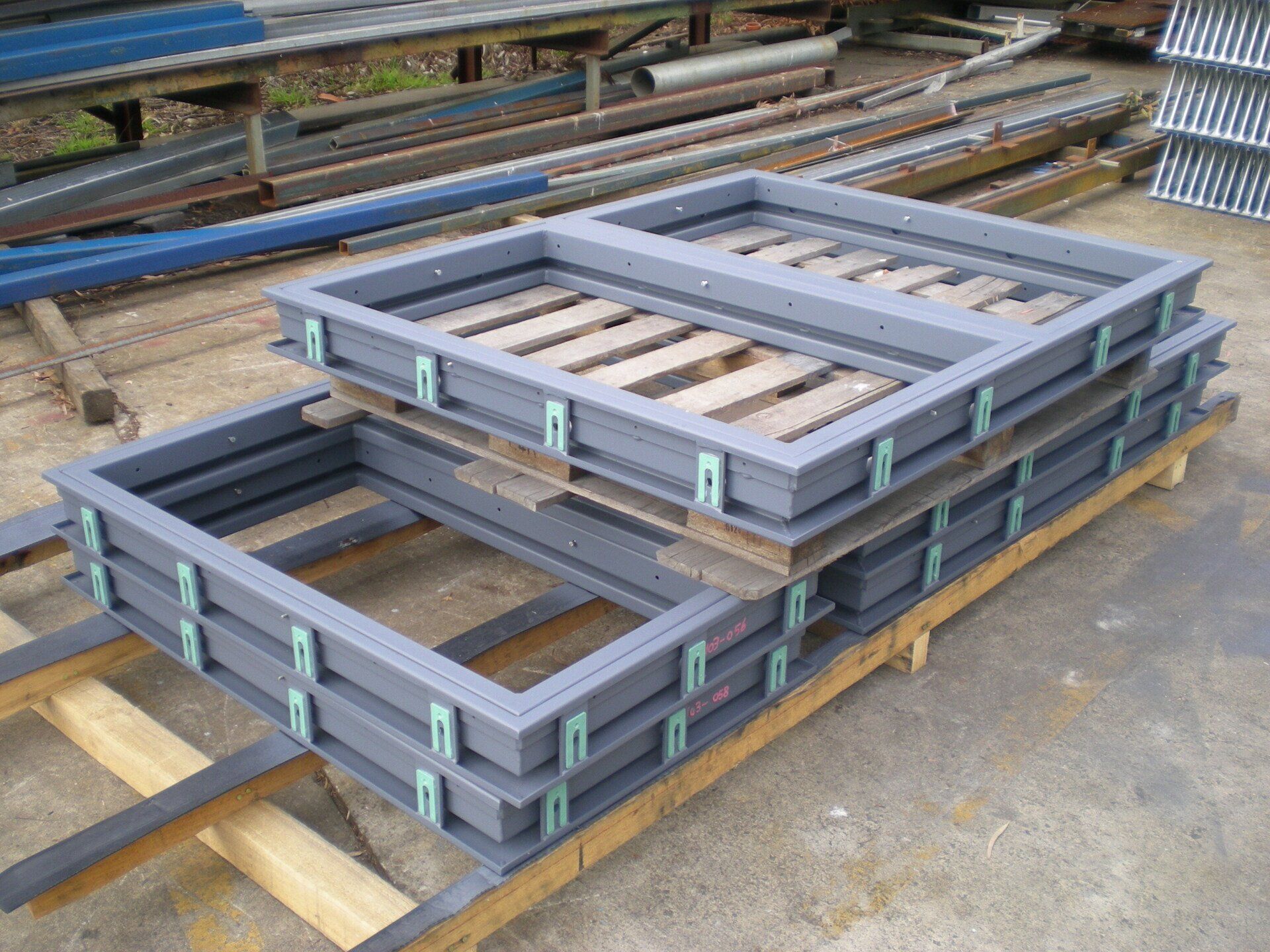 Gray metal frames stacked on wooden pallets, outdoors, with other metal pieces in the background.