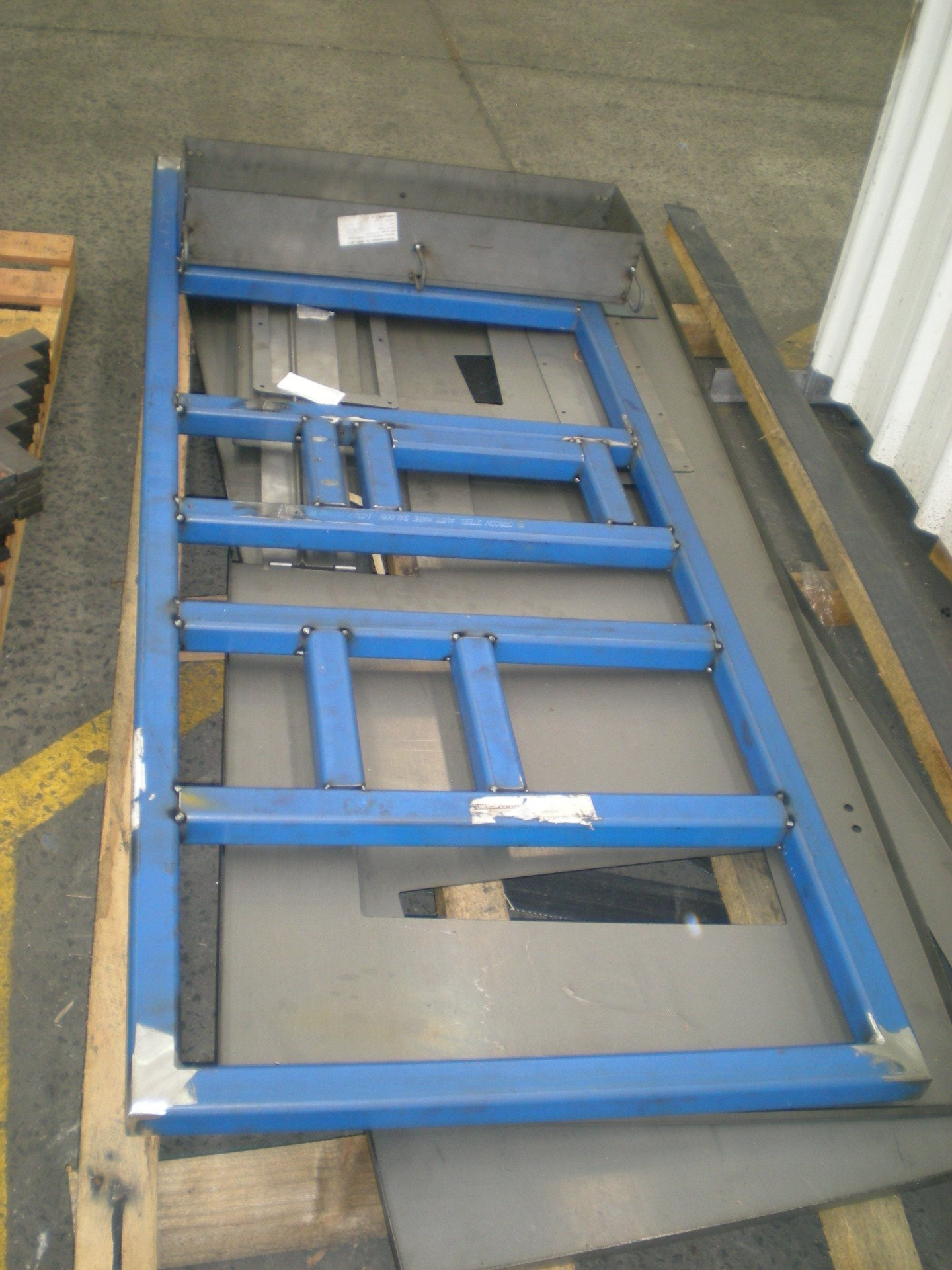 Blue metal frame structure with crossbars, sitting on gray metal sheets, industrial setting.