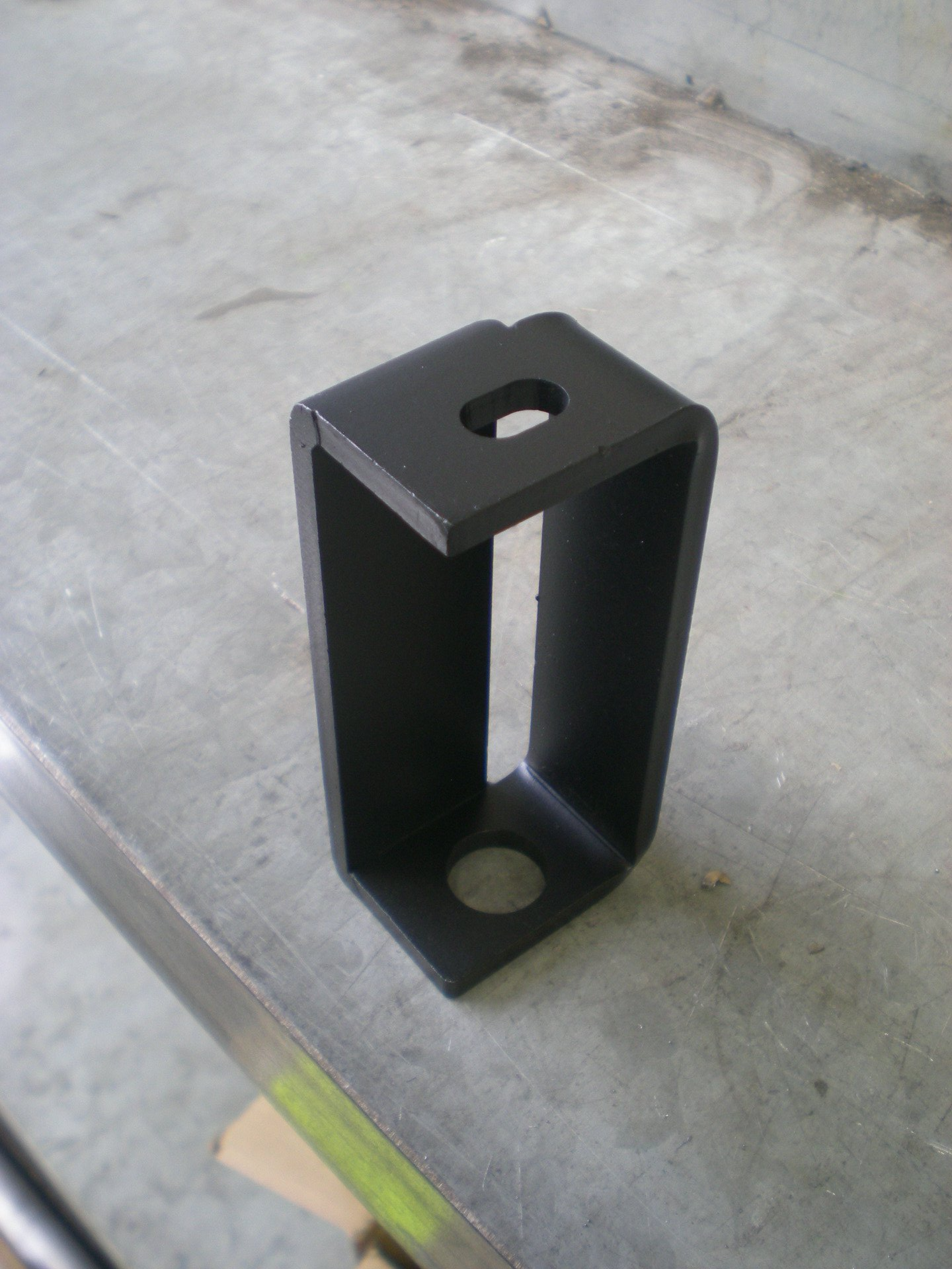 Black, L-shaped metal bracket with a hole in the top and bottom for mounting. It sits on a metal surface.