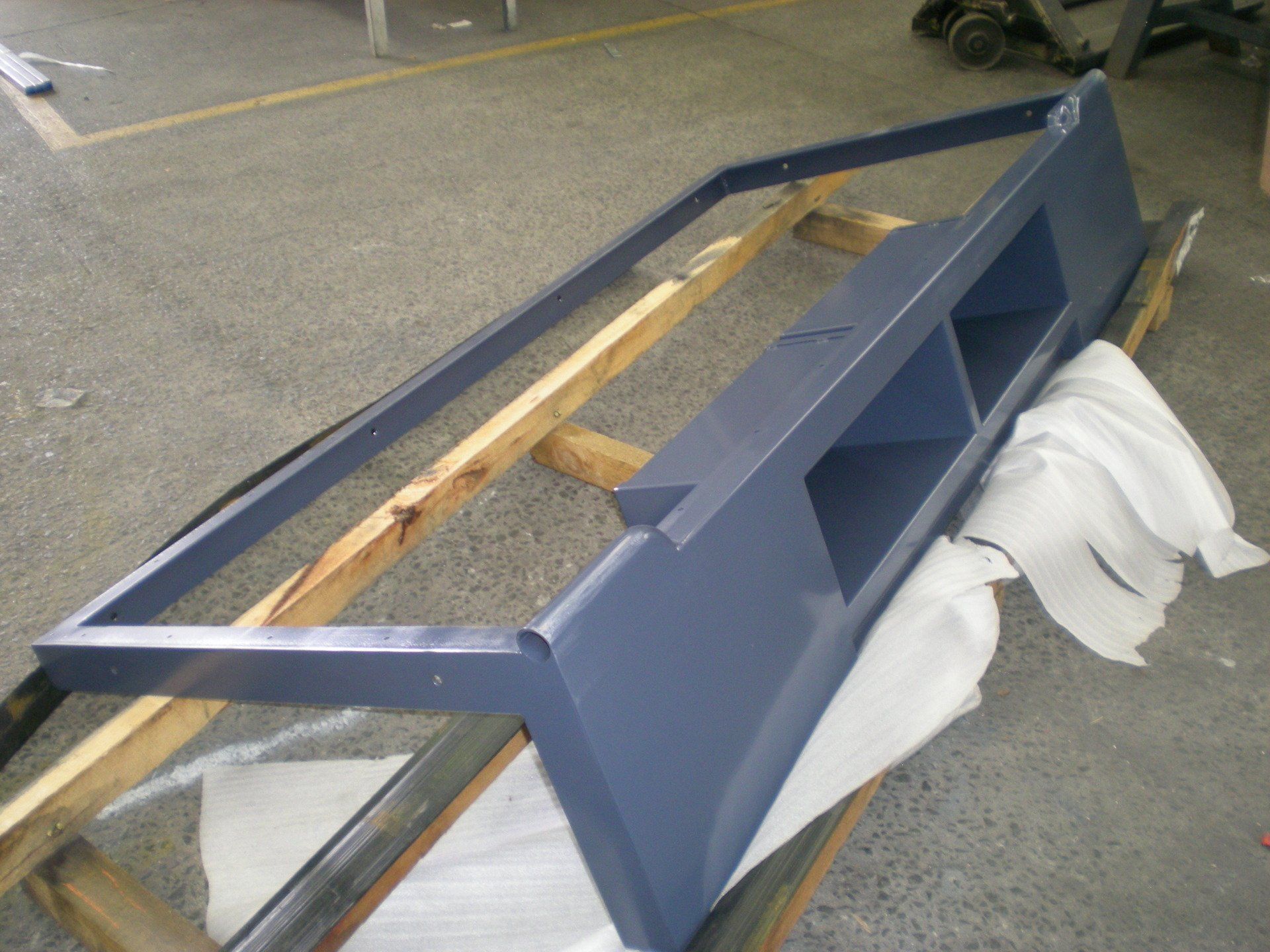 A painted, blue-gray metal frame with three shelves resting on wooden supports on a gray floor.