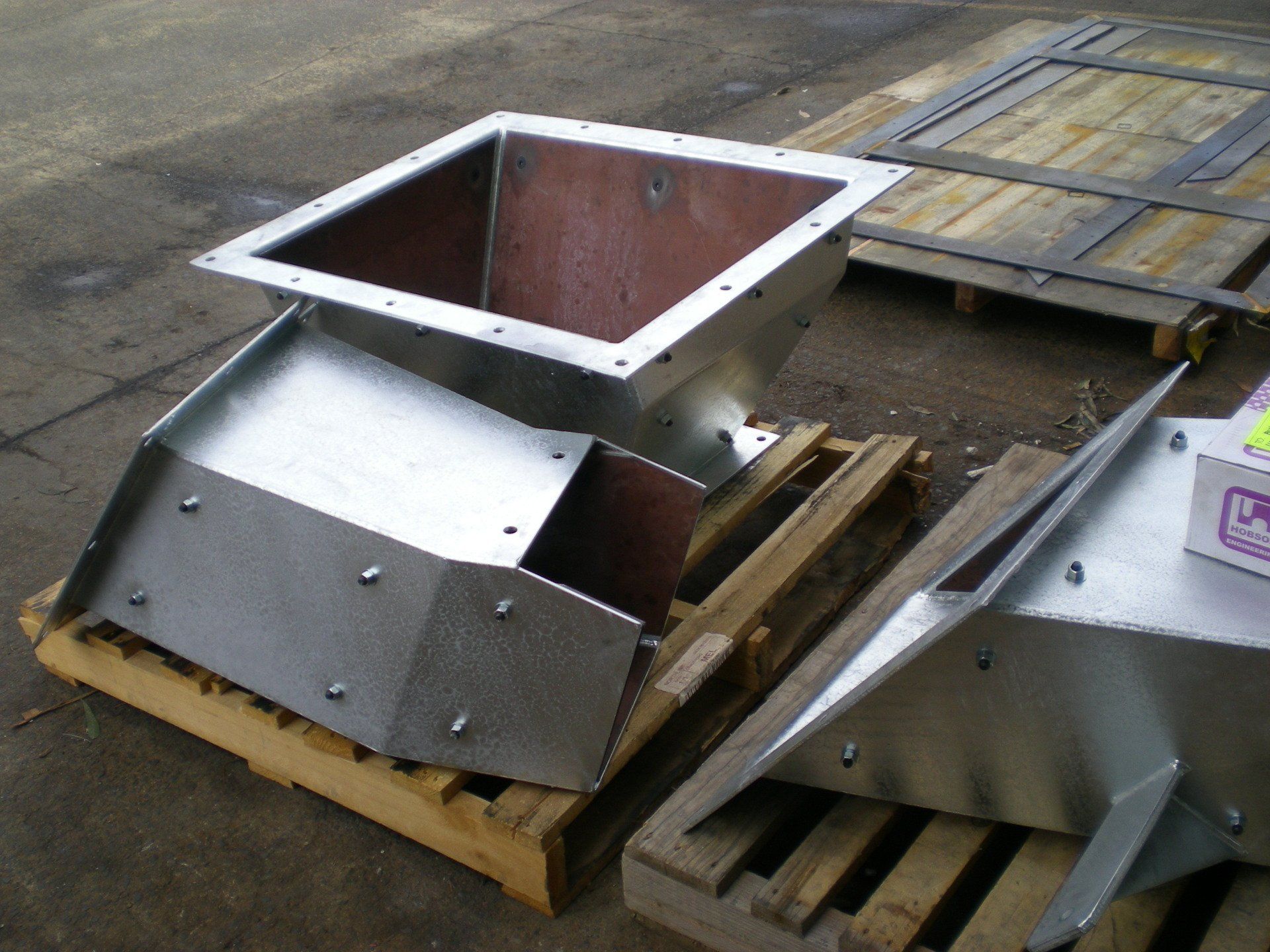 Metal industrial hopper on a wooden pallet, angled with a rectangular top and a sloped chute.
