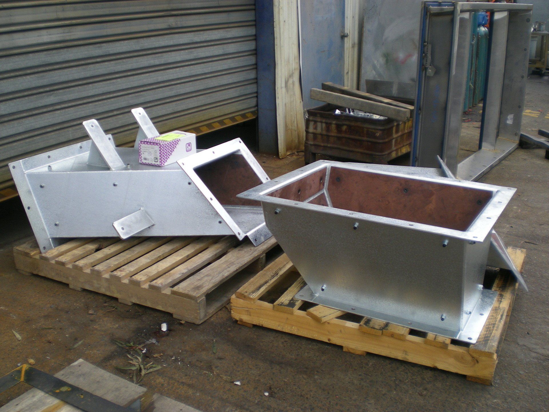 Two metal industrial hoppers on wooden pallets sit in a factory setting. The hoppers are rectangular with angled sides.