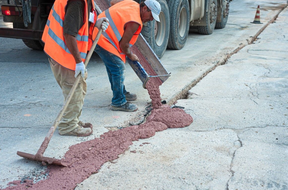 Workers in orange safety vests pour asphalt into a cracked road repair trench