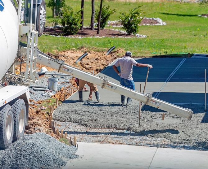 Workers pour concrete at a construction site beside a cement truck and dirt mound.