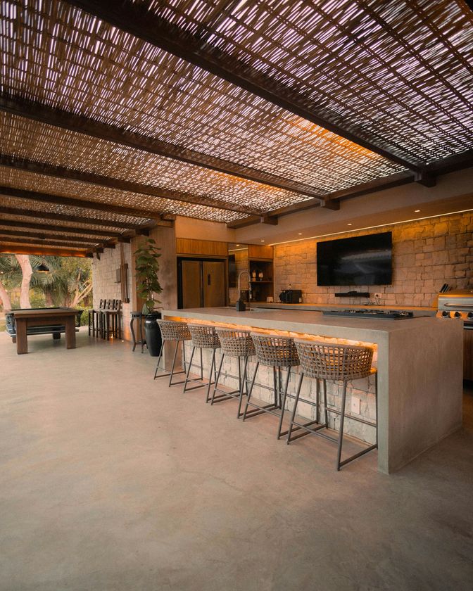 Open-air modern bar with woven roof, concrete counter, stools, and pool table