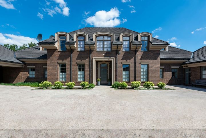 Large brick mansion with arched windows, dormers, and a wide gravel driveway under a blue sky