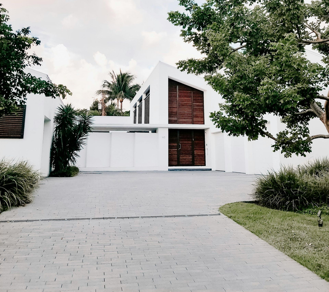 Modern white house with angular design, large wooden shutters, and a paved driveway surrounded by trees.