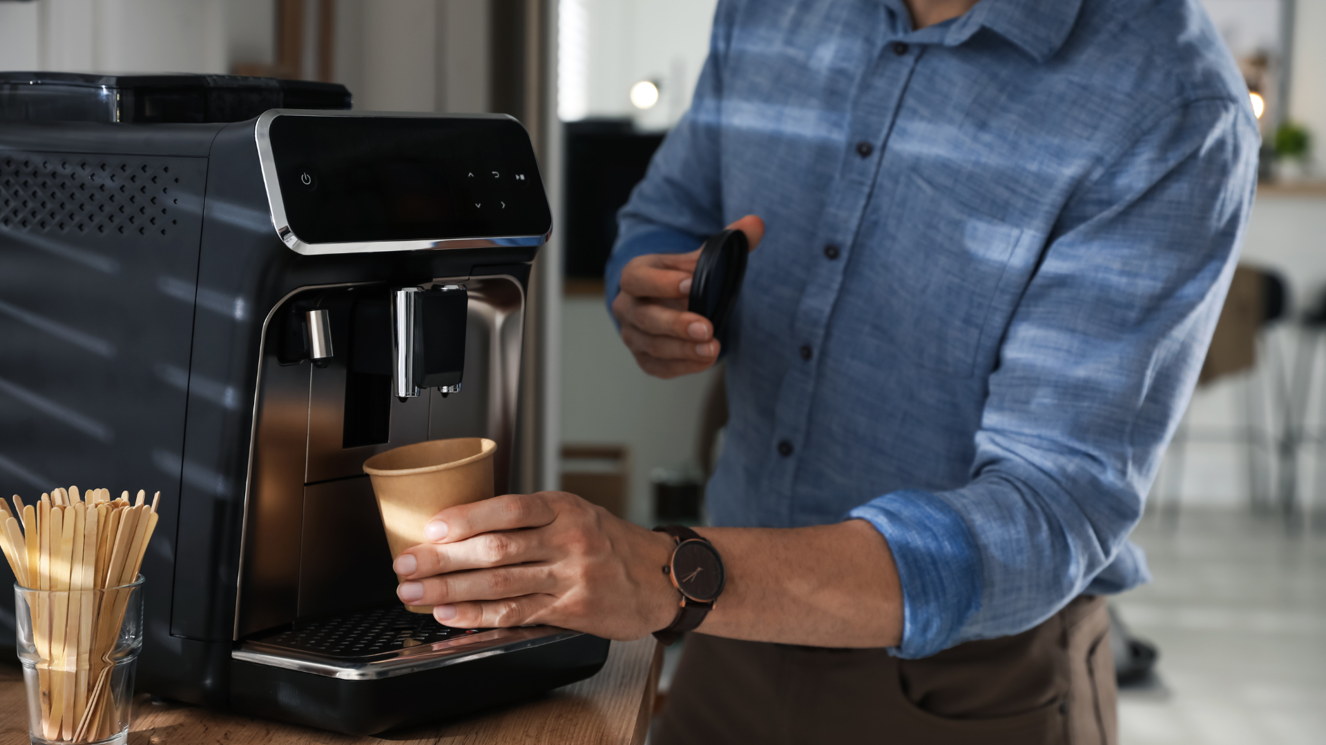 Person placing a paper cup under a black coffee machine to fill it with coffee