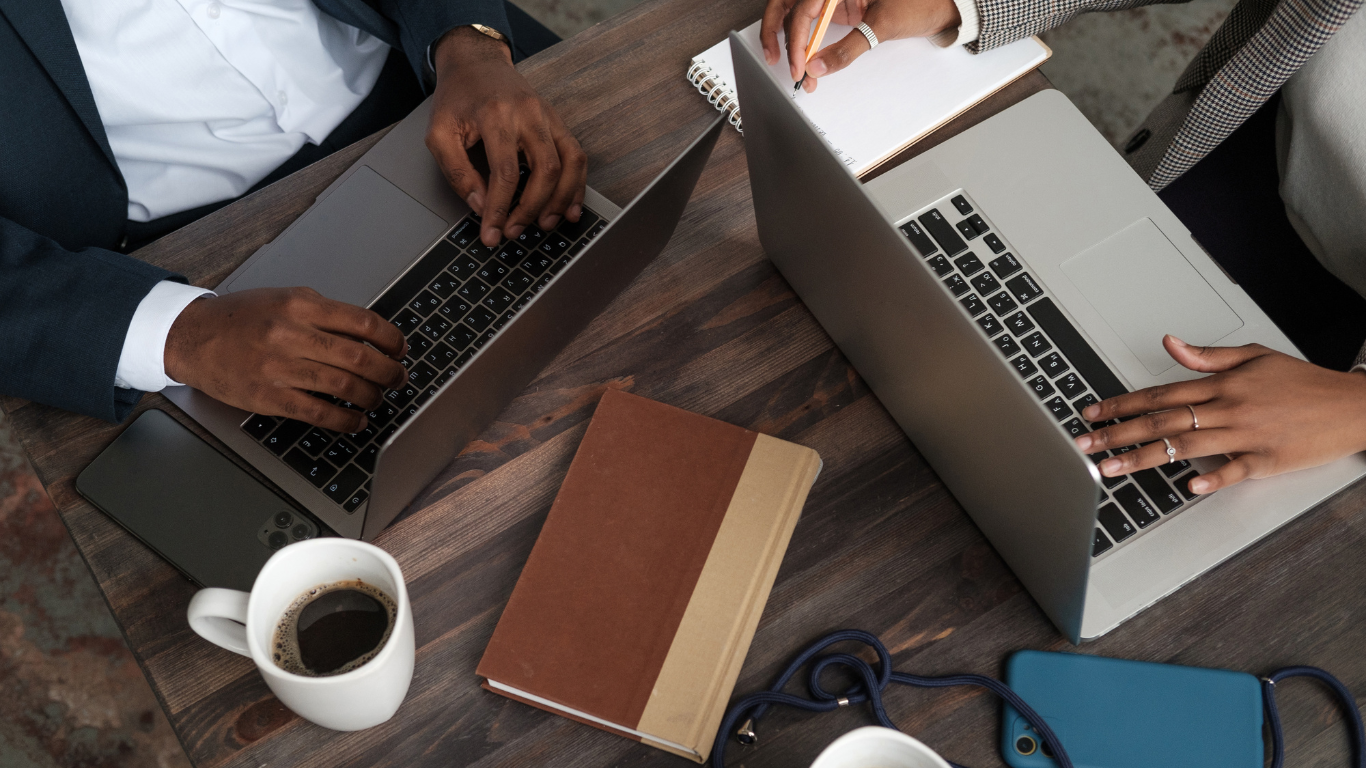 Overhead view of three people working on laptops at a wooden table with coffee and notebooks.