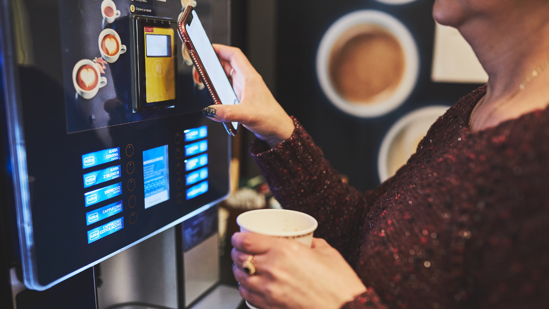 Person using a touchscreen coffee machine while holding a cup, with a coffee cup and display in view