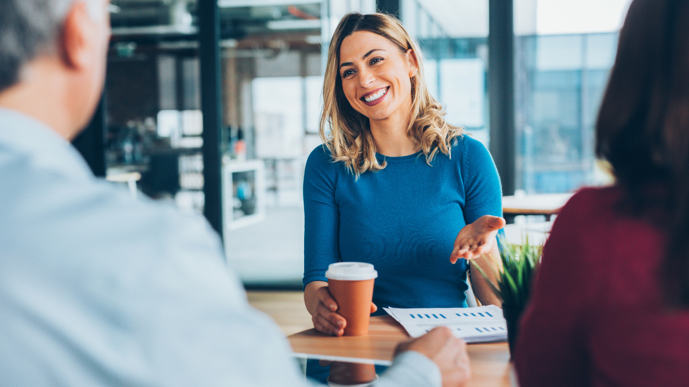 Two people talk across a table with coffee in a bright office meeting space.