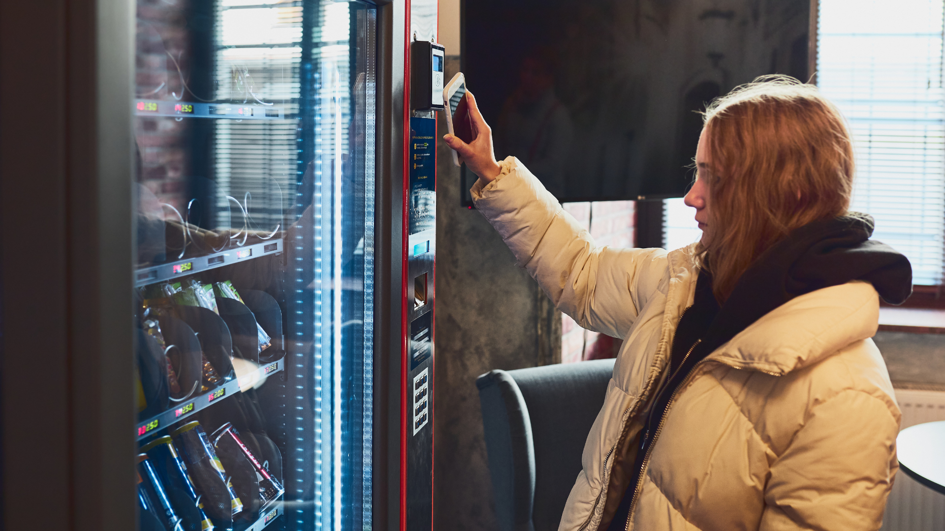 Person in a beige coat reaching toward a vending machine in a dim indoor setting.