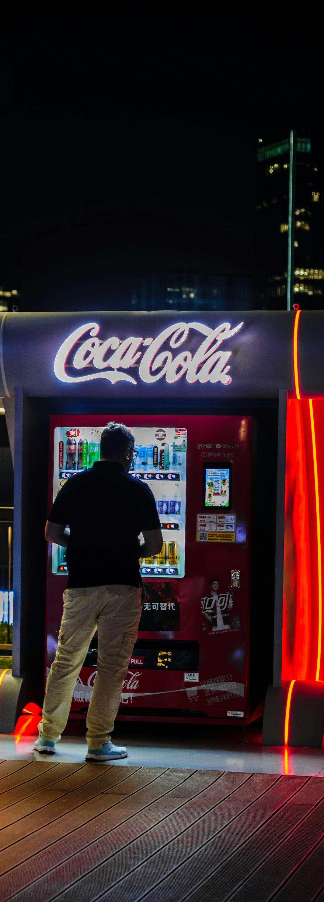 Person using a Coca-Cola vending machine at night, lit by red neon glow in an outdoor setting