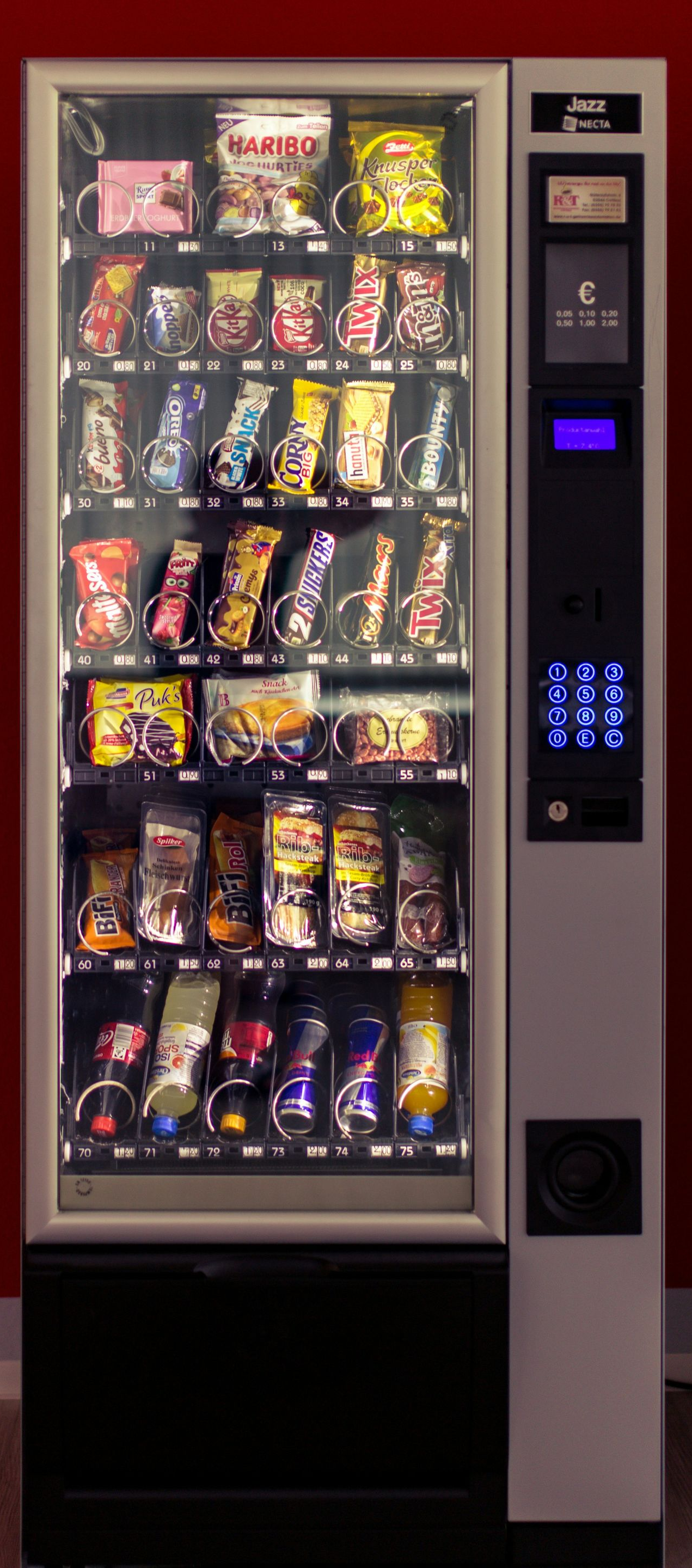 Snack vending machine against a red wall, stocked with chips and candy bars.