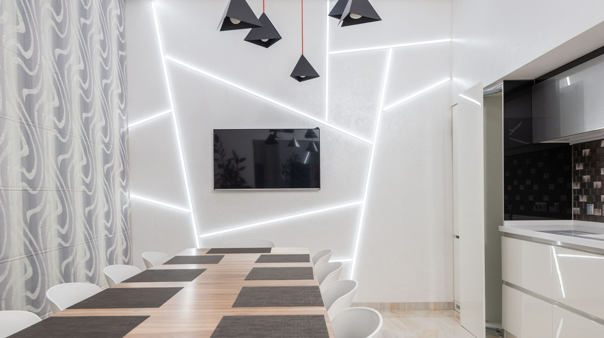 Modern dining room with long table, black chairs, wall TV, geometric lights, and sleek white cabinetry