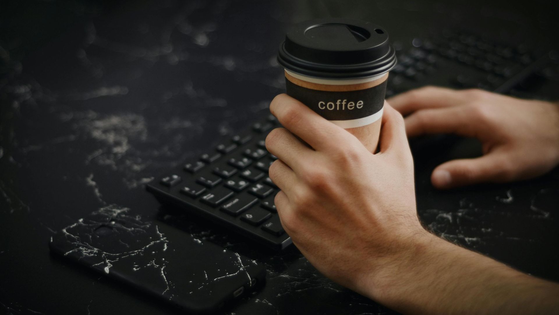 Hands holding a coffee cup beside a keyboard and phone on a dark desk
