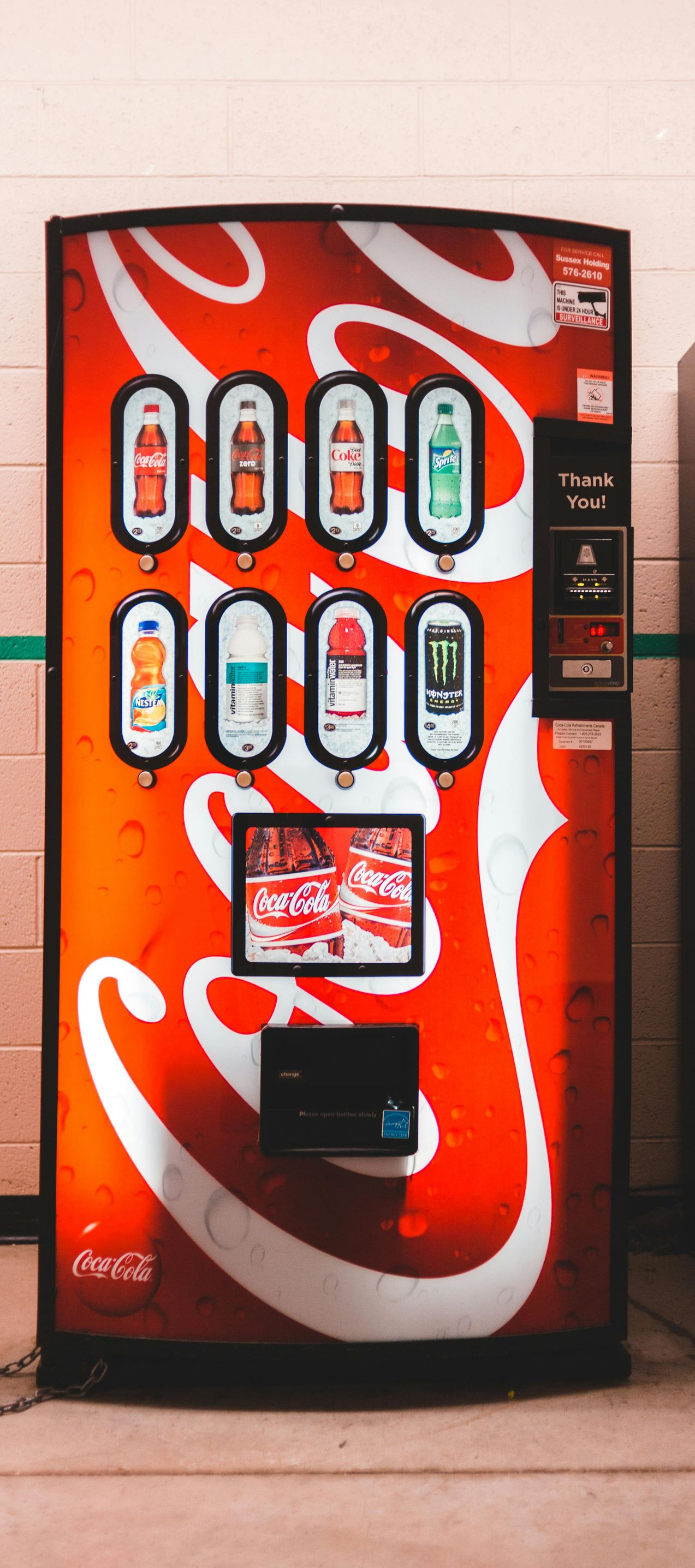 Coca-Cola vending machine with illuminated drink buttons in a hallway, beside a water fountain