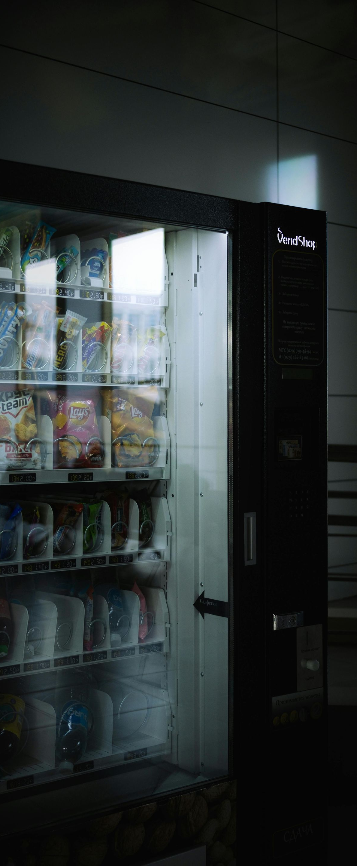 Snack vending machine in a dim hallway with rows of packaged drinks and snacks visible through the glass.