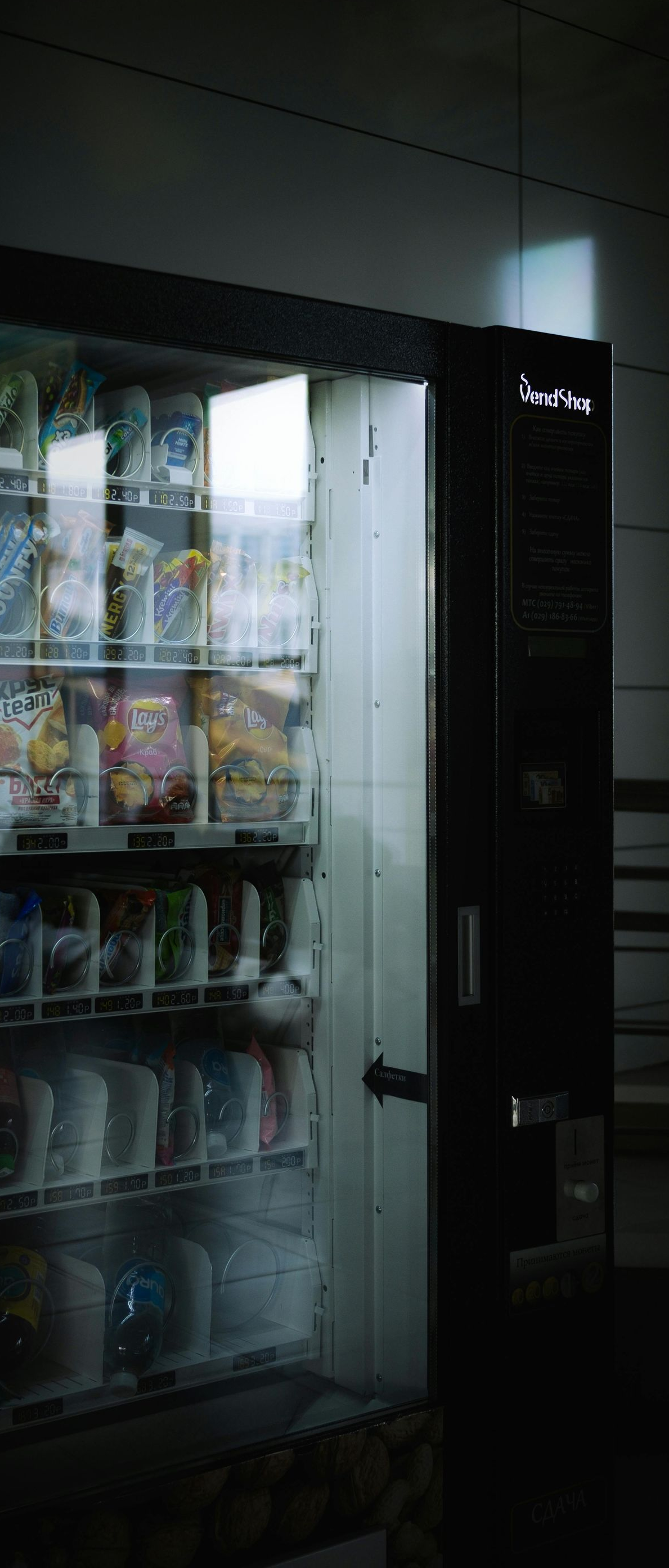 Snack vending machine in a dim hallway with rows of packaged drinks and snacks visible through the glass.