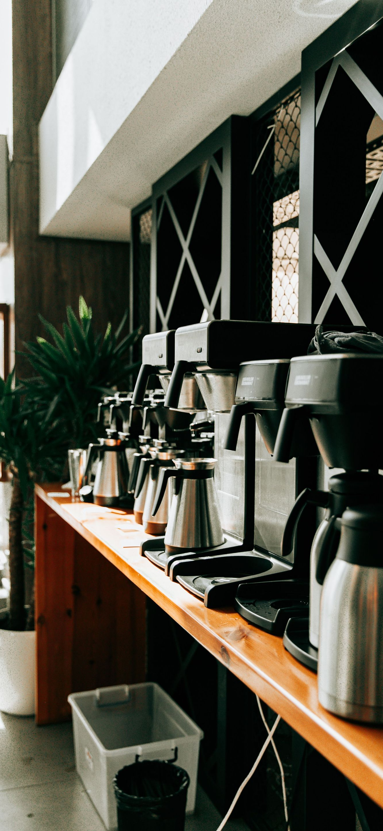Coffee station with several silver brewers and cups on a wooden shelf by a window.