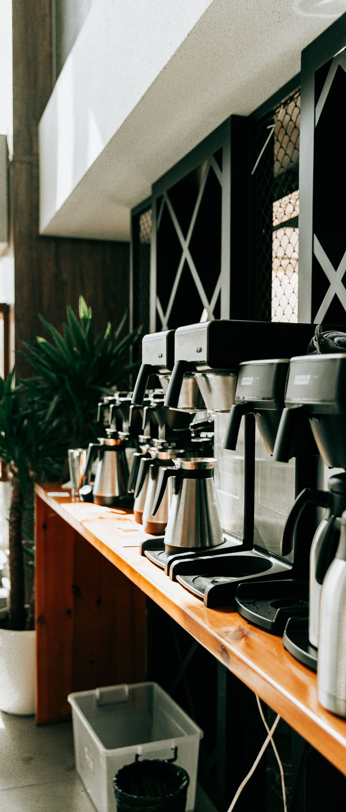 Coffee station with several silver brewers and cups on a wooden shelf by a window.