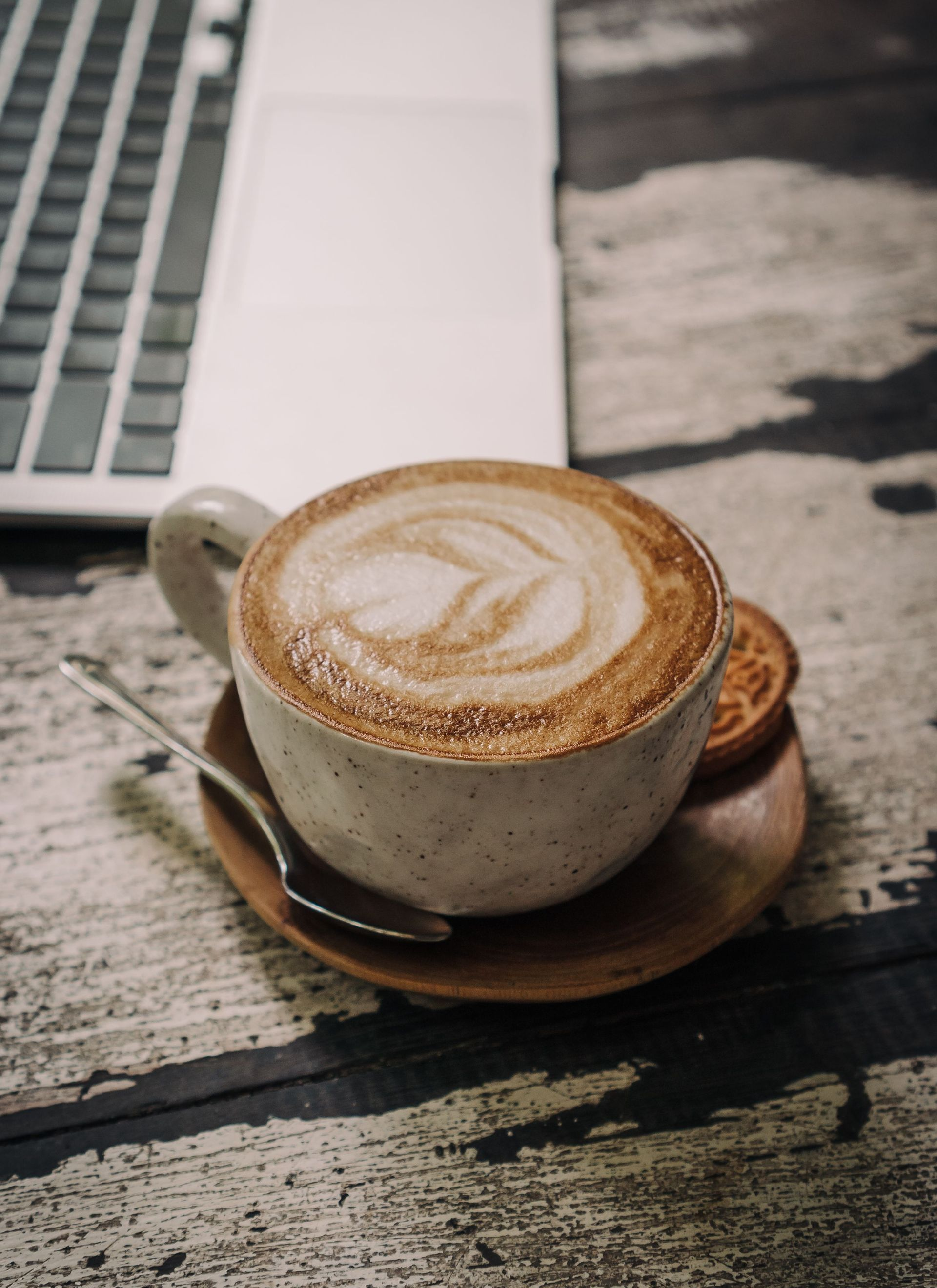 Latte in a beige cup on a saucer beside a laptop on a weathered table