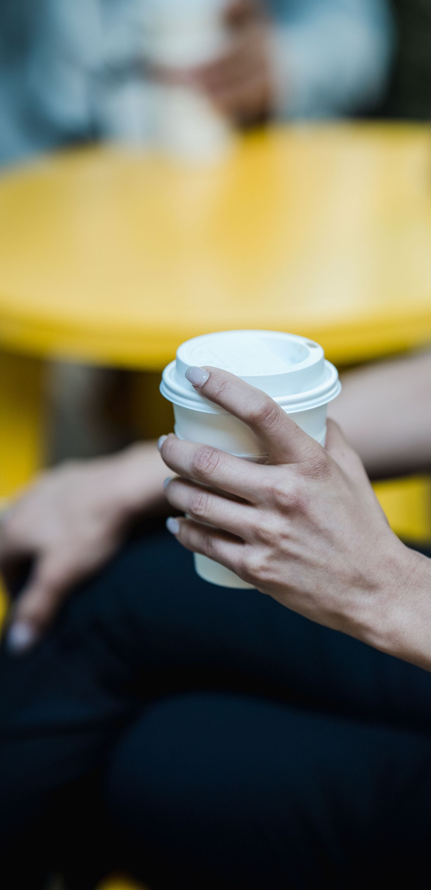 Hand holding a white takeaway coffee cup at a yellow café table, with a blurred background