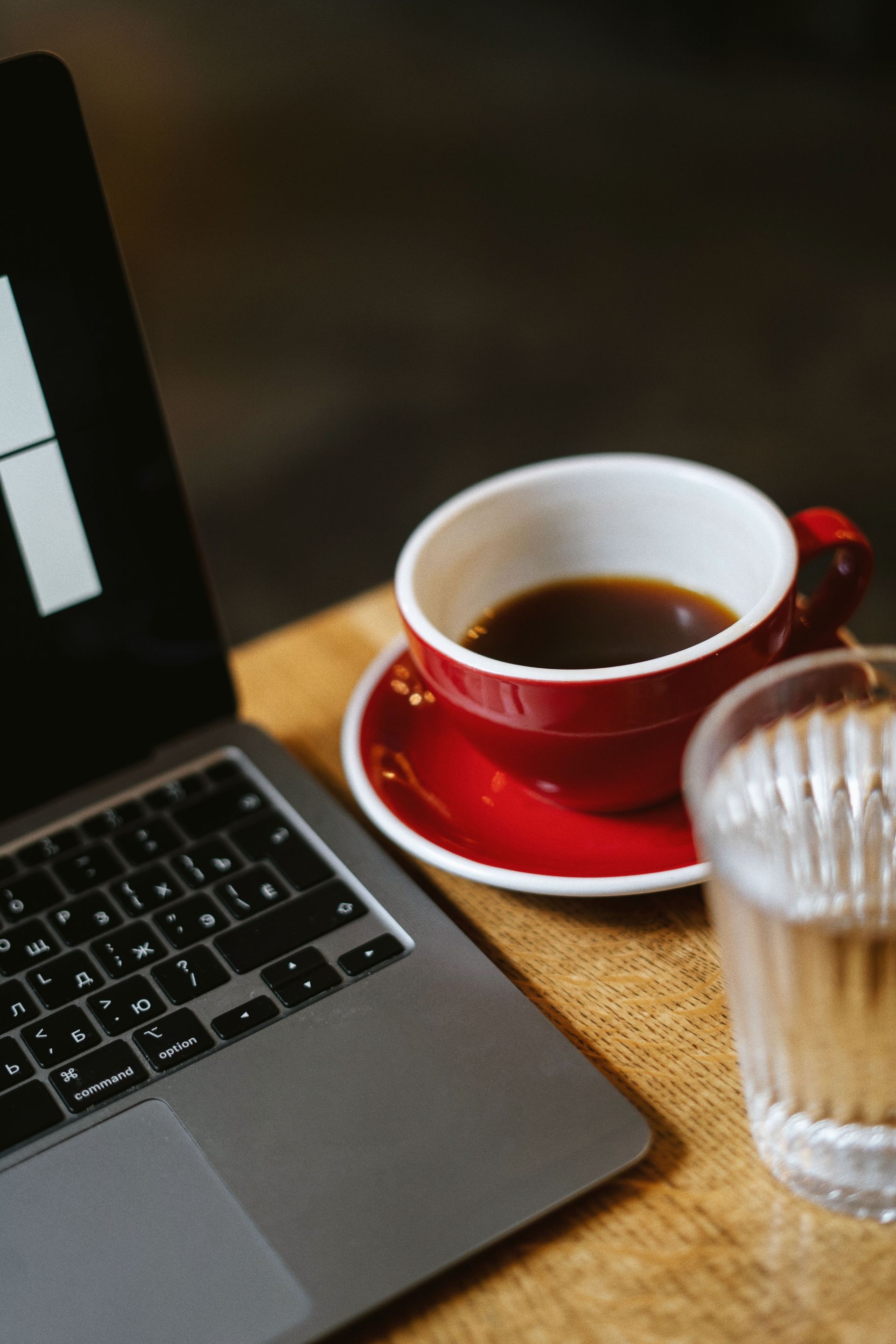 Laptop next to a red cup of coffee on a saucer, with a glass of water on a wooden table.