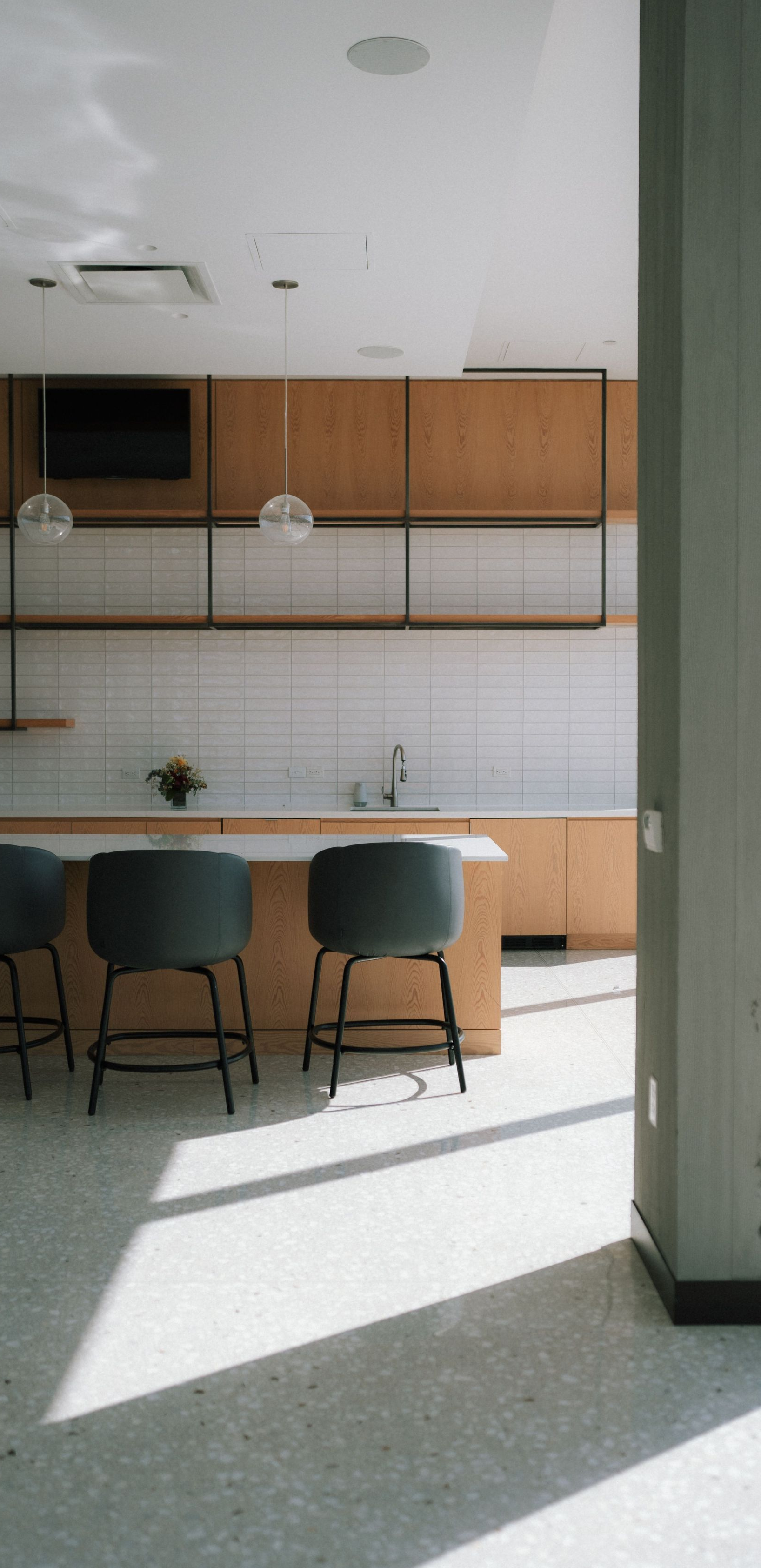 Modern kitchen with wood cabinets, white tile backsplash, and two teal chairs by a sunlit counter