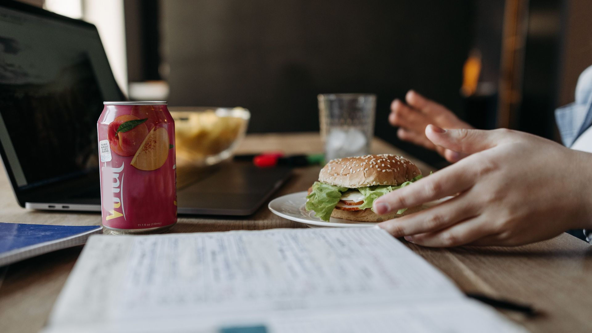 Desk with a burger, soda can, laptop, and hands reaching across a meeting table.