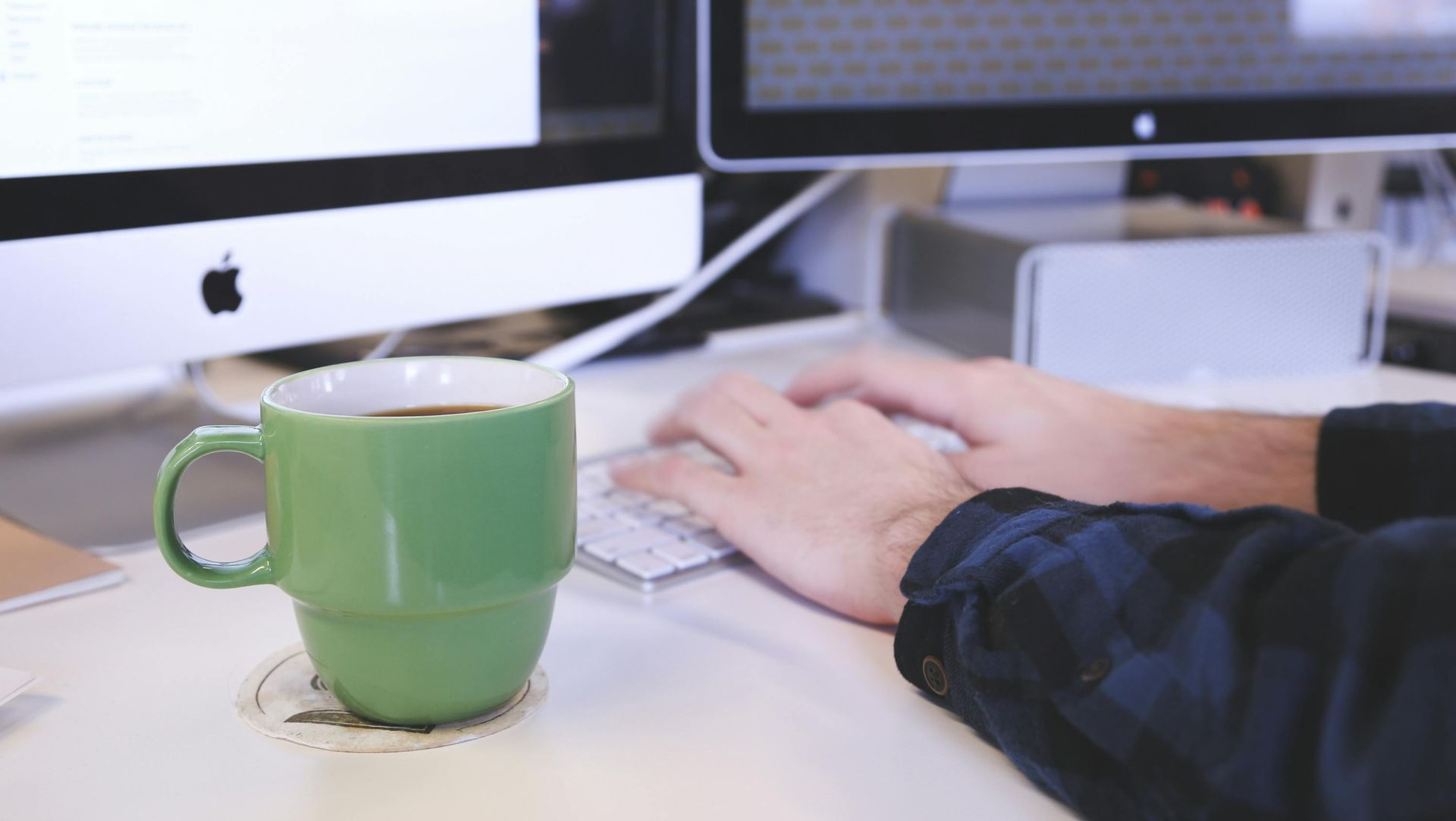 Person typing at an iMac with a green mug on a desk