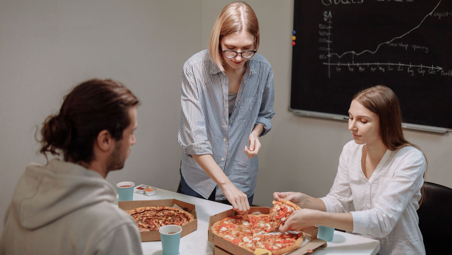 Three people sharing pizza at a table in a meeting room with a goals chart on the wall
