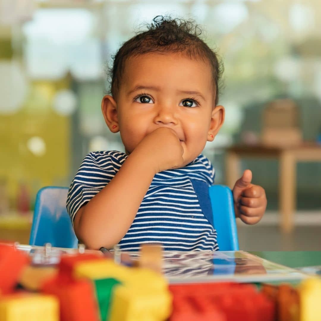Toddler sitting at a table, hand in mouth, with colorful toys in the foreground and a blurred indoor background