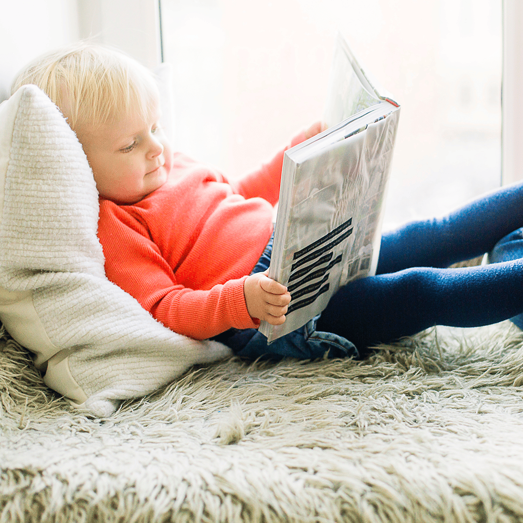 Child in a red sweater reading a newspaper on a white shaggy rug by a pillow