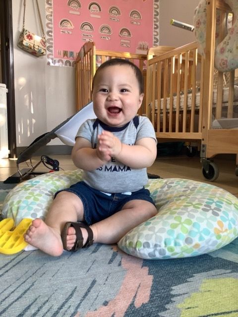 Smiling baby sitting on a patterned cushion in a nursery, clapping hands and wearing a blue shirt.
