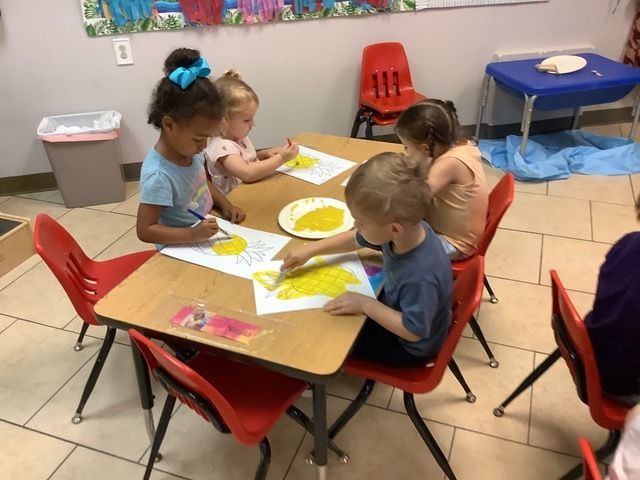 Children painting at a classroom table with yellow paper and brushes, surrounded by red chairs.