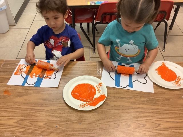 Two children paint orange handprints at a classroom table with paper and paint plates.