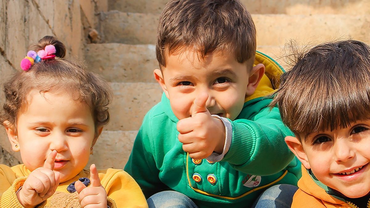 Three children sitting close together, smiling and giving thumbs-up outdoors.