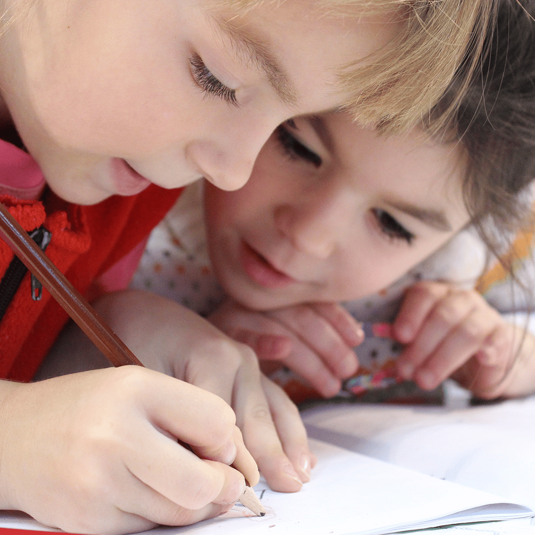 Two children leaning over a sheet of paper, drawing with pencils in close-up.