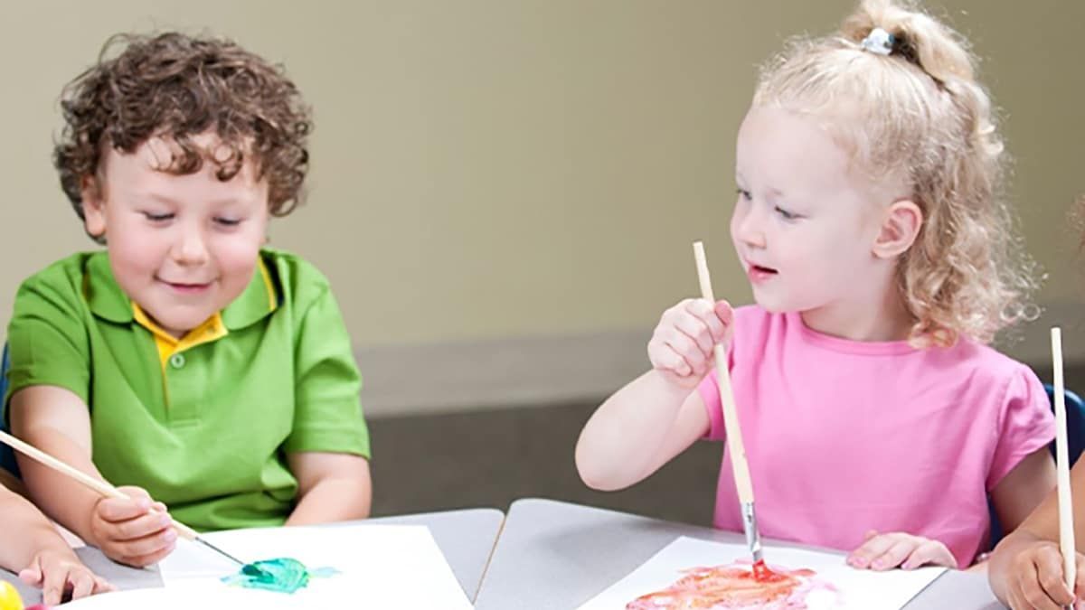 Two children painting at a table, one in a green shirt and one in a pink shirt.