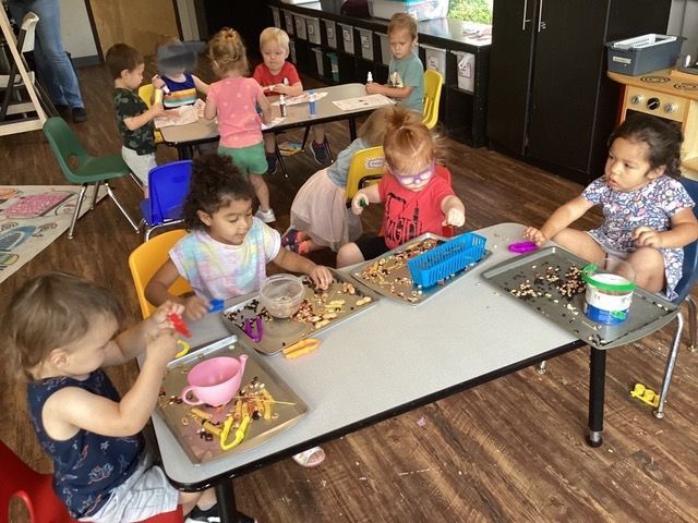 Children doing arts and crafts at tables in a classroom, with beads and trays on the tables