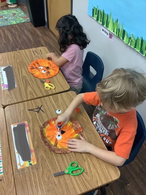 Children painting orange paper pumpkins at a classroom table with scissors and crayons nearby